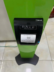 A green stand with a black ZYWELL ticket printer displaying ticket number “010,” used for queue management in service areas.