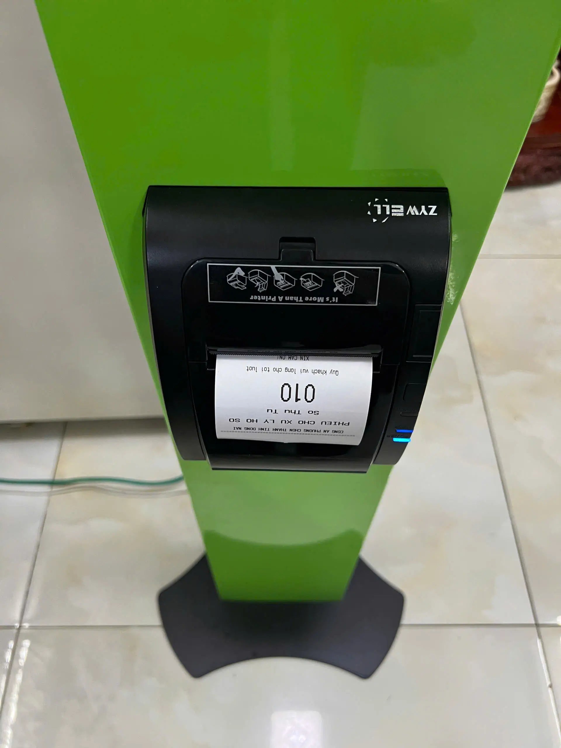 A green stand with a black ZYWELL ticket printer displaying ticket number “010,” used for queue management in service areas.